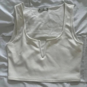 Vit croppad linnetopp från NLY One - Säljer en stilren vit croppad linnetopp från NLY One. Toppen har en djup v-ringning med en snygg detalj framtill och är ärmlös. Perfekt att matcha med höga jeans eller kjol för en trendig look. (Metall) spänne vid v-ringning
