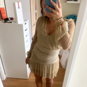 Kort volangklänning i guld - Superfin guldfärgad kort klänning med plisserat tyg, v-ringning och långa transparenta ärmar. Kjolen har volangkant och midjan är markerad med omlottliknande effekt. Är endast använd en gång💓💓I stl xxs/xs från meet x tiger