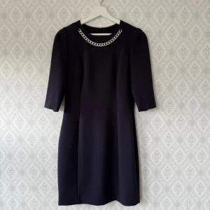 Mörkblå kort klänning med kedjedetalj - Snygg mörkblå kortklänning med halvärmar och en cool silverfärgad kedja vid halsringningen. Klänningen har en rak, stilren passform och dragkedja längs ryggen. Perfekt för dig som gillar stilrena detaljer och vill sticka ut lite extra. Använd 2 gånger. Rökfritt hem!