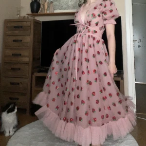 Autentisk "Strawberry dress" av Lirika Matoshi  - Drömmig rosa långklänning från Lirika Matoshi med puffärmar, djup v-ringning och omlottknytning i midjan. Den är äkta, skräddarsydd (storlek M) och köptes för ca 6000 kr året 2020. Ej använd, endast prövad fåtal gånger. Klänningen har glittriga jordgubbsmotiv, volangkant nertill och är gjord i tunn tyll. Perfekt för dig som vill sticka ut med en söt och lekfull look.