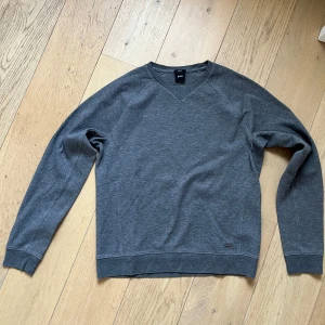 Grå slim fit tröja från Hugo Boss - Snygg grå tröja från Hugo Boss i slim fit-modell. Tröjan har rund halsringning med V-detalj, långa ärmar och diskret metallogo vid nederkanten. Tillverkad i mjuk bomullsmix som känns skön mot huden. Perfekt för en clean och stilren look.