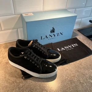 Lanvin Paris (Glansiga) - Snygga svarta Lanvin med vit platt sula och glansig lackad tå. Skorna har snörning och är tillverkade i en mix av mocka och lack, vilket ger en lyxig och stilren vibe. Perfekta för dig som gillar exklusiva detaljer och klassisk design. Storleken på denna är UK7 som motsvarar: 42-43. 🤗