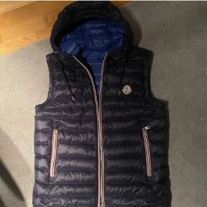 Moncler väst  - Snygg mörkblå dunväst från Moncler med avtagbar huva och blått foder. Västen har två fickor med dragkedja och Monclers logga på bröstet. Röd, vit och blå randig detalj längs dragkedjorna ger en sportig vibe. Den är i nyskick såklart äkta