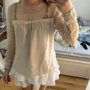 Beige blus med spetsdetaljer - Romantisk beige blus med transparent spets upptill och på ärmarna. Blusen har rund halsringning, långa ärmar med knappar vid manschetten och fina spetskanter. Luftigt material som ger en drömmig känsla och passar perfekt till en söt eller bohemisk stil.
