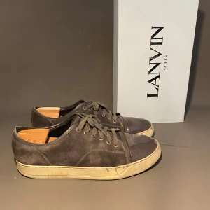 Bruna sneakers från Lanvin i mocka - Snygga bruna sneakers från Lanvin med ovandel i mocka och tåparti i skinn. Skorna har rund tå, snörning och en beige platt sula som ger en clean och klassisk look. Perfekta för dig som gillar stilrena och exklusiva sneakers.