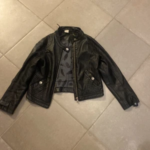 Svart skinnjacka från Lindex - Cool svart skinnjacka från Lindex Kids med snygga detaljer som quiltade partier vid axlar och sidor, dragkedja framtill och tryckknappar vid fickor och ärmslut. Jackan har en klassisk bikerstil och är fodrad med mönstrat tyg.