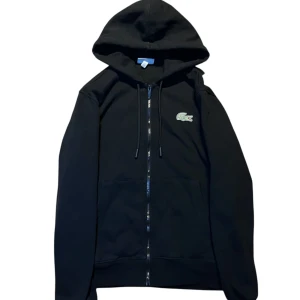 Svart hoodie från Lacoste  - Kvitto Finns!      Svart hoodie från Lacoste med klassisk krokodillogga på bröstet. Tröjan har huva med snörning, hel dragkedja framtill och två fickor. Perfekt för dig som gillar en clean och stilren look.