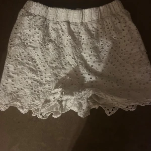 Vita broderade shorts med volangkant - Superfina vita shorts i bomull med broderade detaljer och hålmönster. De har elastisk midja och volangkant nertill för en söt och somrig vibe. Perfekta till varma dagar och lätta att matcha med en enkel topp.