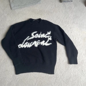 Svart stickad Saint Laurent tröja - Svart stickad tröja från Saint Laurent med vit, fuzzy logga framtill. Klassisk rund halsringning och ribbade muddar vid ärmslut och nederkant. Pris kan diskuteras 