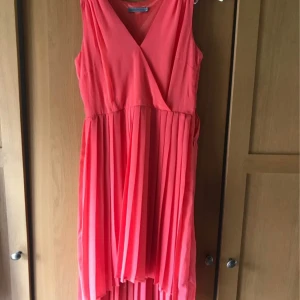 Korallrosa omlottklänning med plisserad kjol - Säljer en snygg korallrosa klänning med omlottliv och v-ringning. Kjolen är plisserad och har en lätt asymmetrisk längd. Ärmlös modell som passar perfekt till varma dagar. Superfin färg som verkligen poppar!