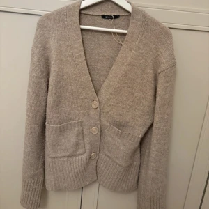 Kofta - Mysig beige kofta från Gina Tricot med djup v-ringning, stora knappar framtill och två praktiska fickor. Tröjan är stickad i ett mjukt material och har långa ärmar samt ribbade muddar och nederkant. Perfekt för lager-på-lager-stil.