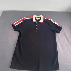 Svart pikétröja från Gucci med röd/vit detalj - Gucci pikétröja i svart med beige krage och röda/vita detaljer längs axlarna. Snygg logga på ärmen och klassisk pikéknäppning framtill. Perfekt för dig som vill sticka ut med en lyxig och sportig vibe. Storlek L men kan passa för även personer med storlek m