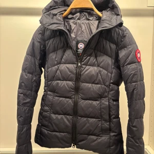 Svart dunjacka från Canada Goose XS - Svart dunjacka från Canada Goose i storlek XS. Jackan har quiltad design, huva med dragsko och dragkedja framtill. Klassisk logga på ärmen och smidig passform som funkar perfekt när det är kallt ute.