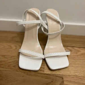 Vita klackskor - Stilrena vita sandaler med smala remmar och fyrkantig tå. Skorna har hög klack och öppen tå, perfekt för att lyfta din outfit. Snygg minimalistisk design med en modern vibe. Tillverkade i syntetmaterial.