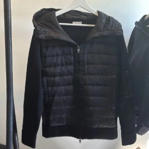 Moncler cardigan - Svart pufferjacka från Moncler med stickade ärmar och quiltad front. Jackan har en stor huva, dragkedja framtill och Moncler-logga på ärmen. Perfekt för dig som vill ha en snygg och sportig look med premiumkänsla.
