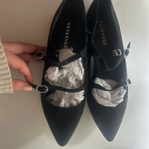 Svarta ballerinaskor från Reserved - Stilrena svarta ballerinaskor från Reserved med spetsig tå och dubbla remmar med silvriga spännen. Skorna är i mocka-look och har en elegant och trendig vibe, perfekta för att lyfta vilken outfit som helst.