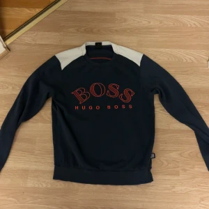 Mörkblå tröja från Hugo Boss - Snygg mörkblå tröja från Hugo Boss med kontrasterande vita axelpartier och röd logga framtill. Tröjan har rund halsringning och långa ärmar, perfekt för dig som gillar stilren design med en sportig touch.