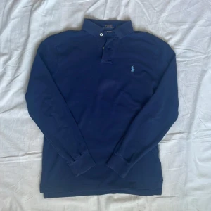 Marinblå långärmad pikétröja Polo Ralph Lauren - Säljer en marinblå långärmad pikétröja från Polo Ralph Lauren. Klassisk krage med två knappar och den ikoniska broderade loggan på bröstet. Tröjan är i bomull och har en normal passform, perfekt till jeans eller chinos.