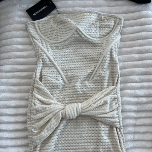 Vit kort klänning från PrettyLittleThing - Supersöt vit kort klänning med strukturerat tyg och rosett framtill. Axelbandslös modell och figurnära passform. Oanvänd med prislapp kvar från PLT. 