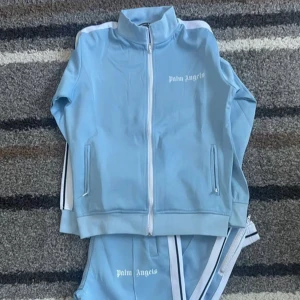 Ljusblå vindjacka från Palm Angels - Snygg ljusblå  Set från Palm Angels med vit dragkedja och vita detaljer längs ärmarna. Jackan har hög krage, två fickor med dragkedja och logga på bröstet. Fick den som present och den vart rätt så stor på mig, både koftan och byxorna är i mycket bra skick och skulle haft kvar den ifall jag hade den storleken som behövdes