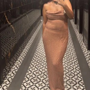 Glittrig champagnefärgad långklänning - Säljer en superfin långklänning i champagnefärg med glittriga detaljer. Klänningen är axelbandslös och har en snygg draperad urringning. Den sitter tight och framhäver figuren, perfekt för en kväll när du vill sticka ut.