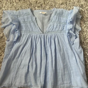 Ljusblå blus med volangärmar - Supersöt ljusblå blus med v-ringning och volangärmar. Blusen har broderade detaljer och rynk vid axlarna, vilket ger en romantisk vibe. STL 158/164!!!