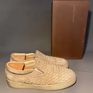 Beige slip-on sneakers från Bottega Veneta - Snygga slip-on sneakers från Bottega Veneta i beige mocka med deras ikoniska flätade mönster. Skorna har en platt sula och rund tå, vilket ger en clean och stilren look. Perfekta för dig som gillar exklusiva och trendiga detaljer.