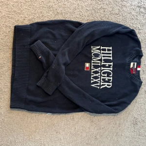 Mörkblå stickad tröja Tommy Hilfiger - Snygg mörkblå stickad tröja från Tommy Hilfiger med stort vitt tryck och logga framtill. Klassisk rund halsringning och ribbade muddar vid ärmslut och nederkant. Perfekt för dig som gillar stilren och tidlös design.