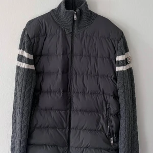 Moncler Cardigan  - Säljer en grå pufferjacka från Moncler med stickade ärmar och rygg i kabelstickat mönster. Jackan har dragkedja framtill, höga ribbade muddar och vita ränder på ärmarna. Perfekt för dig som vill ha en unik och snygg jacka med sportig vibe.