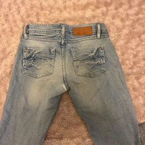 Ljusblå bootcut jeans med broderade fickor - Säljer ett par ljusblå jeans med bootcut-modell och snygga broderade detaljer på bakfickorna. Jeansen har låg midja och är sjukt snygga. Säljer för att de tyvärr inte passar mig längre:( de har ett håll på baksidan som jag har sytt igen men det är inget som syns när man har de på sig!