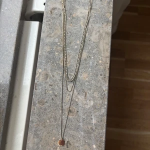 Snyggt lager-på-lager halsband i silver - Trendigt halsband i silverfärg med två kedjor i olika längder. Den ena kedjan är tunn och den andra har en lite grövre länk. Längst ner hänger en rund, slät berlock som ger en stilren och modern känsla. Perfekt för att lyfta vilken outfit som helst.