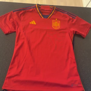 Spansk landslagströja Adidas M - Röd fotbollströja från Adidas för Spaniens landslag. Klassisk design med gula och blå detaljer, broderat landslagsmärke och Adidas-logga på bröstet. V-ringad krage med ränder i rött, gult och blått. Tillverkad i lätt och ventilerande material. Använd 1 gång