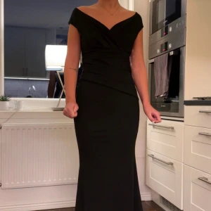Svart långklänning med v-ringning - Fin svart långklänning från bubbleroom med v-ringad omlott överdel och offshoulder. Klänningen har en figurnära passform och är perfekt för dig som vill ha en elegant och stilren klänning, den passar till fest, bal och allt vad man kan tänka sig använda den till. Använd bara en gång. 