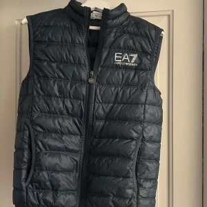 Mörkgrön dunväst från Emporio Armani - Snygg mörkgrön dunväst från Emporio Armani EA7 med quiltad design och dragkedja framtill. Västen har hög krage och två fickor med dragkedja. Perfekt för lager-på-lager och har en stilren logga på bröstet.