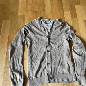 Beige kofta från Pier One - Snygg beige kofta från Pier One med v-ringning och knappar framtill. Långärmad modell i mjukt material, perfekt för lager-på-lager. Klassisk och enkel design som funkar till många olika stilar.