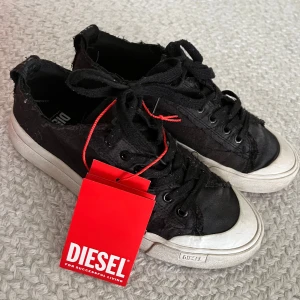 Svarta sneakers från Diesel stl 40 - Använda, men fortfarande fint skick. Från web: These women's trainers flaunt a low-cut satin upper, distressed with frayed raw-cut edges for a broken-in feel. Their tonal design is continued with a matching dirty effect on the sole, extending into a rubber toe cap for added support. Red logo accents sign off the look. Lace-up closure; Rubberised toe guard; Gripped rubber sole; Logo pull tab 100% Polyester Gender: Womens