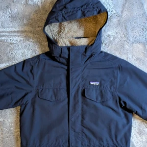 Patagonia teddyfodrad jacka - Jacka från Patagonia i storlek S, med mysigt teddyfoder på insidan och huva. Yttermaterialet är slitstarkt och jackan har två bröstfickor med lock. Perfekt för kalla dagar när du vill hålla stilen och värmen.