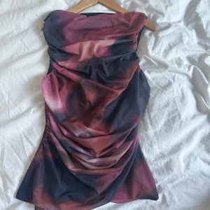 Tubtopp i vinröd och svart mesh - Säljer en oanvänd snygg axelbandslös tubtopp med draperad mesh i vinröda och svarta toner. Toppen har en cool, asymmetrisk form och är figurnära med snygga rynkade detaljer framtill. Säljer den pga jag ej kan returnera för jag klippte bort prislappen innan jag såg den var för liten