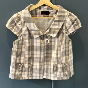 Rutig väst från Zoul med krage - Kort väst från Zoul i rutigt mönster med grå, beige, vit och lila toner. Västen har stor krage, en knapp framtill och två små fickor. Materialet är mjukt och känns som bomull, perfekt för att ge outfiten en preppy touch.