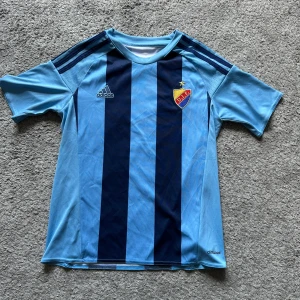 Djurgården fotbollströja Adidas blå - Snygg Djurgården fotbollströja från Adidas med breda ljusblå och marinblå ränder. Klassisk DIF-logga på bröstet och Adidas-logga i blå. Tröjan har korta ärmar och är tillverkad i lätt och ventilerande climacool-material.