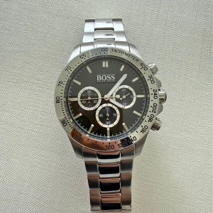 Silverfärgad kronografklocka från Hugo Boss - Stilren kronografklocka från Hugo Boss med svart urtavla, silverfärgade detaljer och robust länkarmband i metall. Klockan har tre mindre urtavlor, tachymeter och tydliga indexmarkeringar. Perfekt accessoar för dig som gillar en klassisk och modern look.                               endast använd vid enstaka tillfällen mycket fint skick 44mm