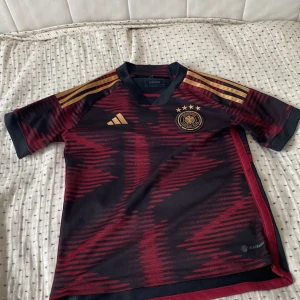 Tysklands bortaställ Adidas 2022 vm  - Snygg Tyskland fotbollströja från Adidas i svart och mörkröd med guldiga detaljer och emblem. Tröjan har korta ärmar, V-ringning och klassiska Adidas-ränder på axlarna. Tillverkad i lätt och ventilerande material, perfekt för match eller träning.