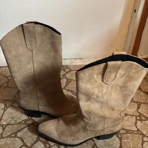 Beiga cowboy boots i mocka - Snygga beiga cowboy boots i mocka med klassiska broderade detaljer och spetsig tå. De har låg klack och svarta sulor, perfekt för dig som vill ha western-vibe i din stil. Lätta att dra på med öglor upptill.