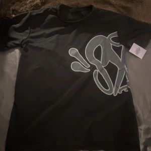 Syna world svart t-shirt  - Säljer en svart Syna World t-shirt med en stor, stilren grafisk print i grått över framsidan. T-shirten har klassisk passform och rund halsringning. Köp t-shirten separat eller tillsammans med det blåa settet. Kom privat för diskussion av pris