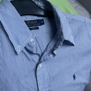 Blårandig skjorta Polo Ralph Lauren - Klassisk blårandig skjorta från Polo Ralph Lauren i slim fit-modell. Skjortan har korta ärmar, knappar framtill och den ikoniska broderade loggan på bröstet. Tillverkad i bomull och passar perfekt för en clean och stilren look.