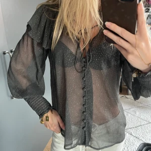 Svart glittrig blus från Zara - Supersnygg svart transparent blus från Zara med glittriga detaljer. Blusen har volang vid axlarna, knytning i halsen, v-ringning och knappar framtill. Långa puffärmar med smock vid ärmslut. .
