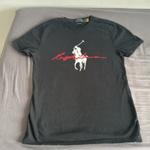 Svart Ralph Lauren t-shirt slim fit - Svart t-shirt från Polo Ralph Lauren i slim fit med klassiskt tryck av polospelare och röd signatur på bröstet. Gjord i mjuk bomull och har korta ärmar. Perfekt för dig som gillar stilrena och ikoniska plagg med en sportig touch.