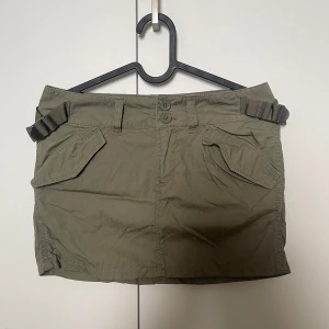 Kortkjol - Snygg olivgrön utility-inspirerad kortkjol, dubbla knappar och spänndetaljer i midjan. Lågmidjad 