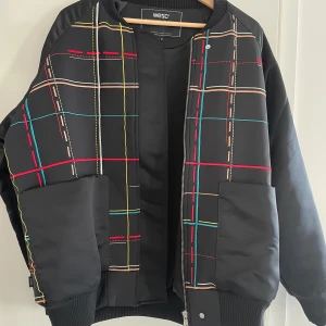 Svart bomberjacka från WESC, rutig - Svart bomberjacka från WESC med broderat rutmönster i rött, blått, gult, beige och vitt. Jackan har ribbade muddar, dragkedja och två stora fickor framtill. Snygg och unik design som sticker ut med sina färgglada linjer.