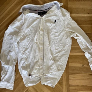 Vit skjorta från Ralph Lauren - Klassisk vit skjorta från Ralph Lauren med broderad logga på bröstet. Skjortan har lång ärm, knappar framtill och en tidlös krage. Perfekt för dig som gillar stilrena och ikoniska plagg. Materialet känns mjukt och är troligtvis bomull.